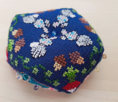 a photo of a biscornu, taken at an angle, with dark blue fabric and cross stitch designs of acorns, rabbits and radishes around the edges